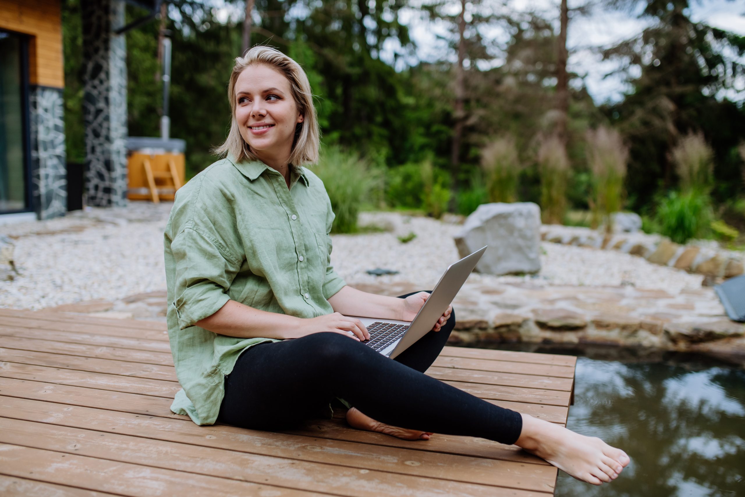 Woman, freelancer working on laptop, sitting on a pier by the backyard lake, a concept of remote office, work during vacation.