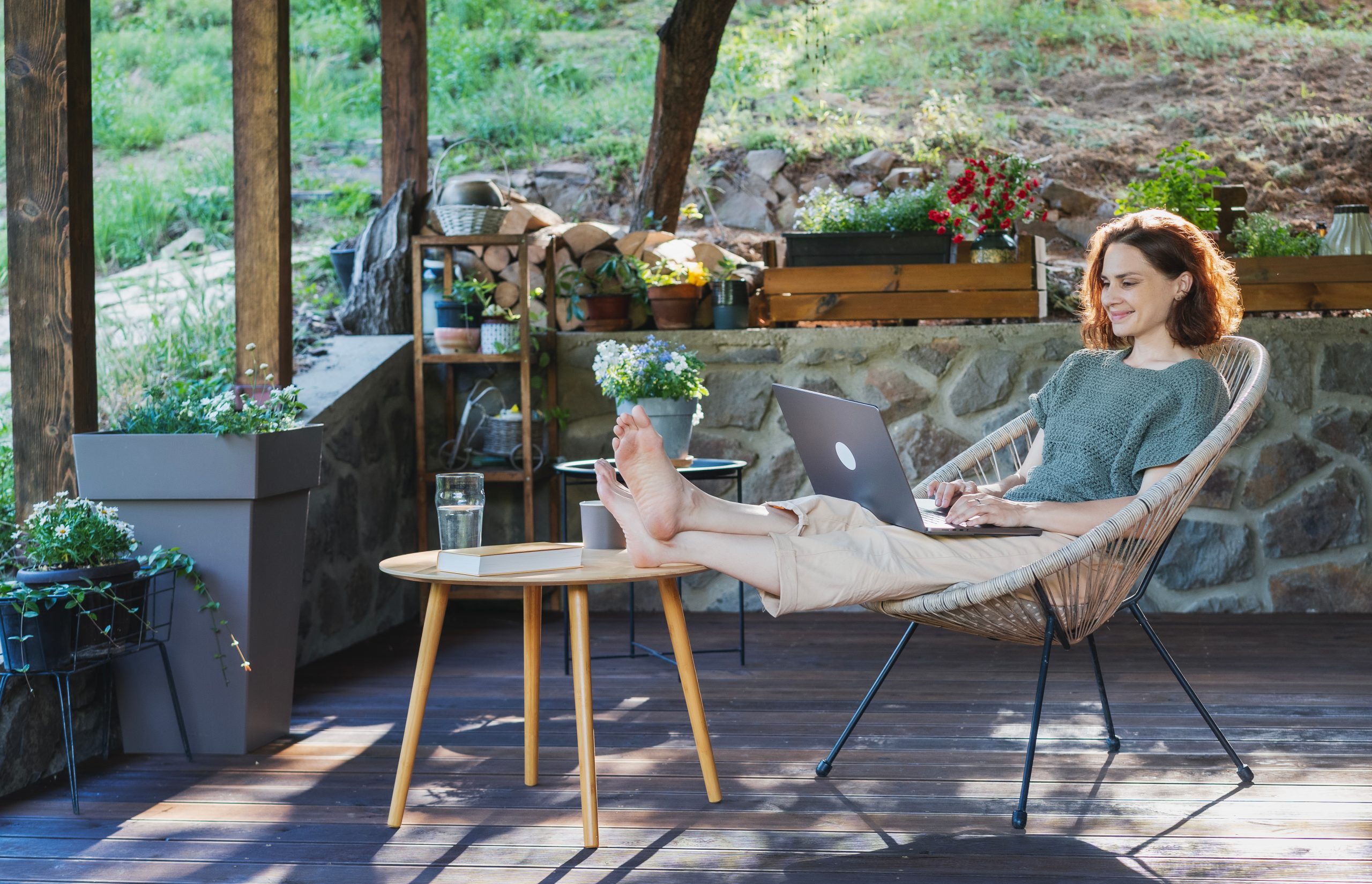 Young adult Caucasian woman sitting on cottage terrace using laptop in summertime, online work